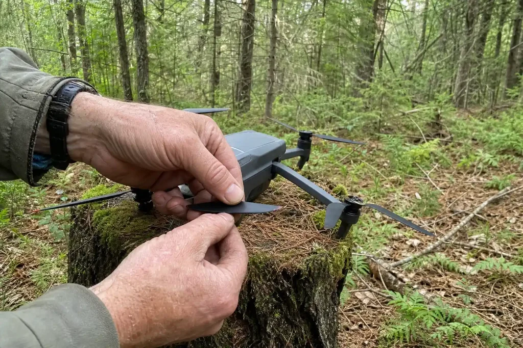 Väderbitna händer justerar propellern på en jordbruksdrönare för gödsling med drönare, vilande på en mossig stubbe i skogen.