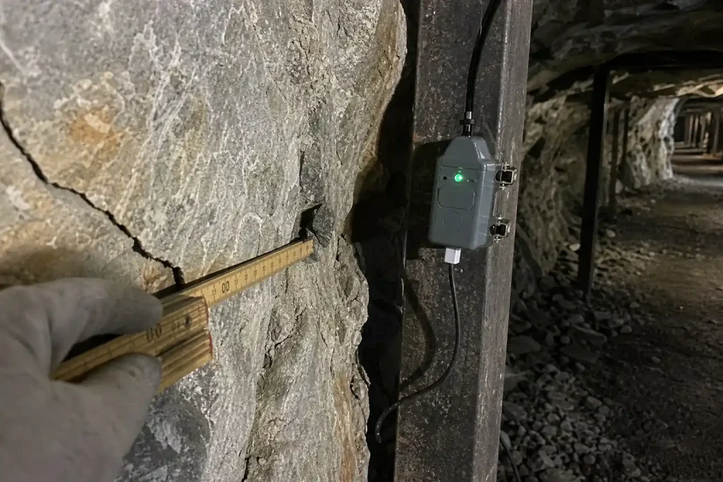En handskbekklädd hand mäter en bergsspricka med tumstock bredvid en AI-sensor i en gruvtunnel.
